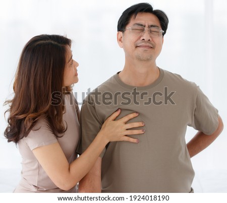 Portrait shot of senior Asian lover couple wearing eyeglasses. Husband suddenly feels uncomfortable from acute back pain with worried wife taking care her husband with isolated white background