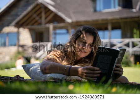 Young latin female smiling reading book outdoors on a pillow with a vegetation and house background