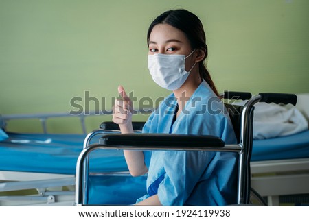 Portrait of a smiling Asian young female patient with surgical mask sitting on wheelchair in hospital room showing thumbs up gesture and sign to camera