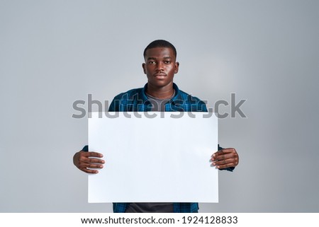 Serious young african american guy in casual clothes looking at camera, displaying blank banner ad, holding it in front of him, posing isolated over gray background. Front view