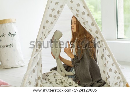 A girl with a soft toy hare sits in a children's toy wigwam, a child is at home in a hut, a girl plays with toys.
