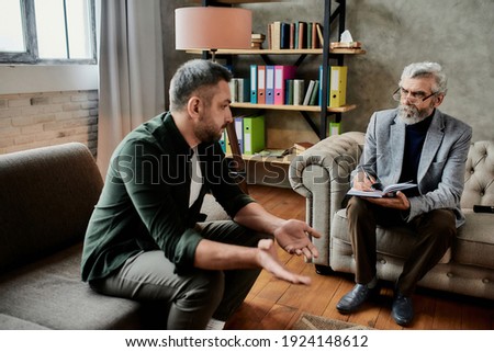 Angry young caucasian man talking about his problems with psychotherapist during appointment in office, selective focus. Psychotherapy concept Royalty-Free Stock Photo #1924148612