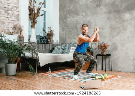 A young man goes in for sports at home, online workout from the laptop. The athlete makes squat , watches a movie  in the bedroom, in the background there is a bed, a vase, a carpet. Royalty-Free Stock Photo #1924186265
