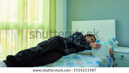 A handsome Caucasian man with long hair lying in his bed and reading a book in a bedroom