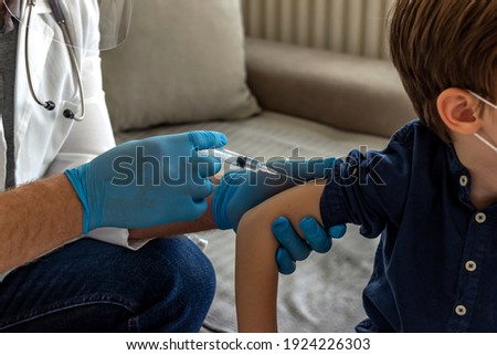 Professional doctor in blue sterile gloves holding syringe and making injection to a little boy in medical mask. Selective focus of boy getting flu shot. Immunisation, medicine and healthcare concept.