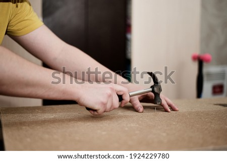A man hammers a nail into a chipboard board with a hammer. Assemble the cabinet at home.