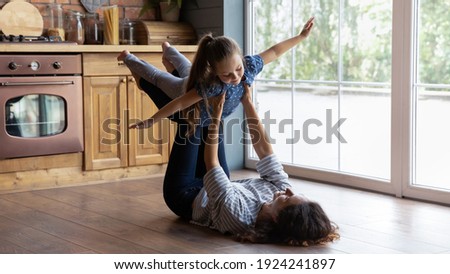 Caring mother holding little daughter pretending flying with hands outstretched, lying on warm wooden floor in kitchen, loving young mum carrying adorable girl, family engaged in funny activity Royalty-Free Stock Photo #1924241897