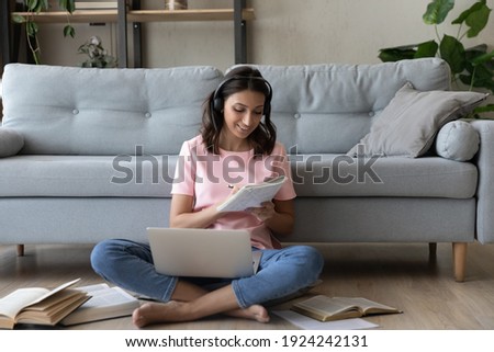 Confident indian woman in headset translate document using web app printed books making notes to paper copybook. Arab female student sit on warm floor at home prepare work write up data from pc screen