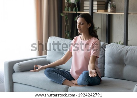 Concentrated young indian female sitting on sofa with crossed legs practicing yoga pranayama exercises. Calm relaxed millennial arabic woman meditating at home taking lotus position relieving stress Royalty-Free Stock Photo #1924242791