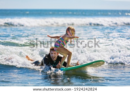 Little surf girl - young surfer learn to ride on surfboard with instructor at surfing school. Active family lifestyle, kids water sport lessons, swimming activity in summer camp. Vacation with child. Royalty-Free Stock Photo #1924242824