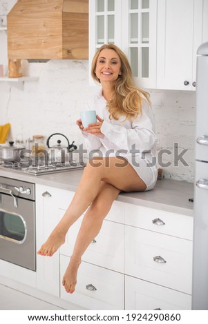 Fun mood. Cheerful beautiful adult woman barefoot sitting on table at home in kitchen