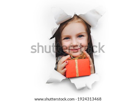 A fanny little caucasian smiling girl looks out of a hole in white pastel paper and holds a red box with a gift in her hand. Christmas and New Year concept and birthday concept. Copy space.