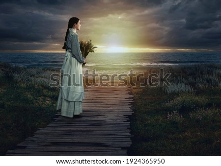 Young victorian girl holding dried flowers in white dress and blue striped blouse on a boardwalk at the coast at sunset.