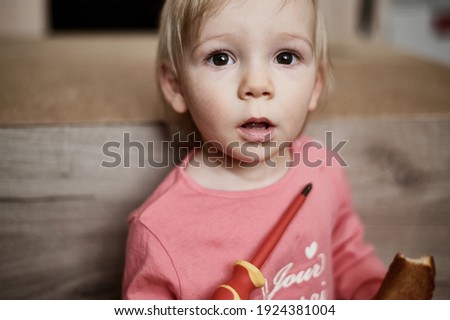 A small child looks into the frame against the background of bokeh with a screwdriver in his hands.