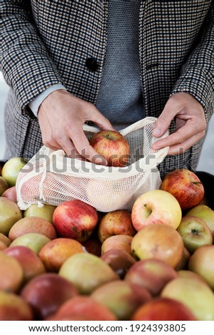 a young caucasian man, wearing a brown coat, uses a textile reusable mesh bag to buy some apples at a greengrocer or a street market, as a measure to reduce plastic pollution