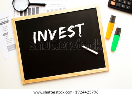 Text INVEST written in chalk on a slate board. Office desk with office supplies. Business concept.