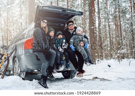 Young family dressed in warm clothing pose on camera sitting on car trunk in winter forest in daytime.