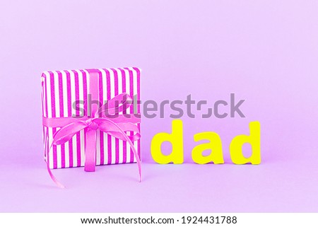 Happy Father's day. Gift box with a ribbon and a bow, the word "dad" from wooden letters on a light purple background. Copy space. Close-up.