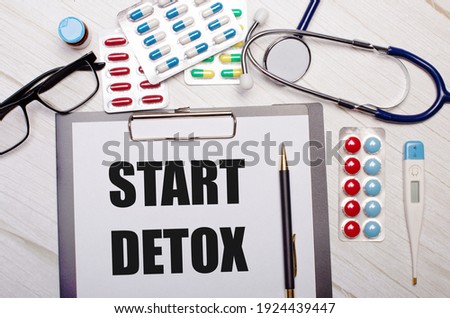 On a light wooden background there is paper with the inscription START DETOX, a stethoscope, colorful pills, glasses and a pen. Medical concept