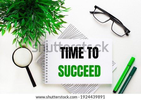 On a white background lies a notebook with the words TIME TO SUCCEED, glasses, a magnifying glass, green markers and a green plant
