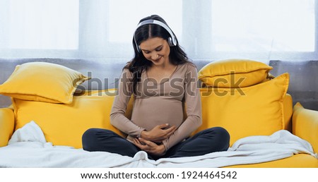 Smiling pregnant woman in headphones embracing belly in living room