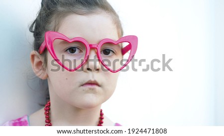 Serious toddler brown eyes girl in red heart glasses looks at camera white background. Symbol of love and Valentines Day