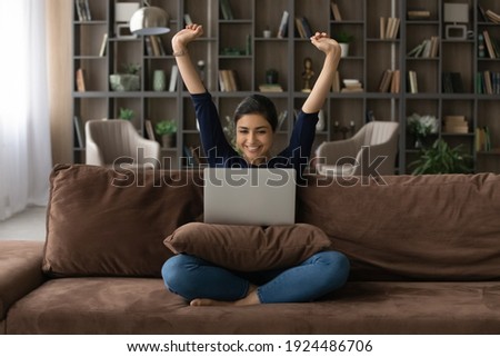 Overjoyed millennial Indian woman sit on sofa in living room feel euphoric read good news on computer. Happy excited young mixed race female triumph with online lottery win or work finish on laptop. Royalty-Free Stock Photo #1924486706