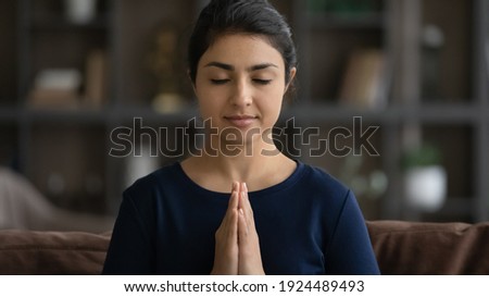 Crop close up of calm Indian woman with eyes closed hold hands in prayer meditate at home. Young mixed race female believer pray to God, feel superstitious religious. Faith, religion concept.