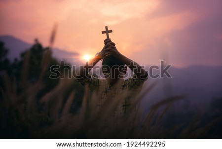 Boy standing holding a Cross overhead for pray to God with light sunset background, beliefs of children concept.