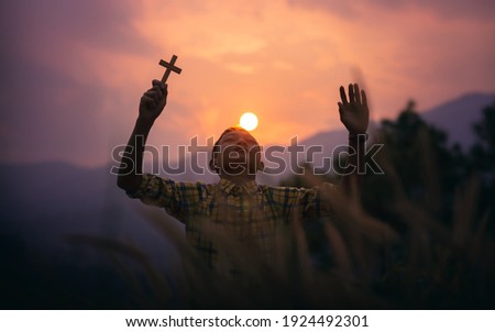 Boy holding a Cross overhead for pray to God with light sunset background, beliefs of children concept.