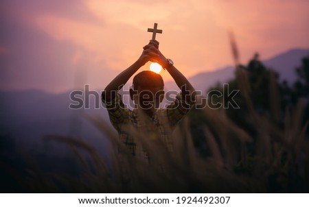 Christian boy standing holding a Cross overhead for pray and worship to God with light sunset background, beliefs of children concept.