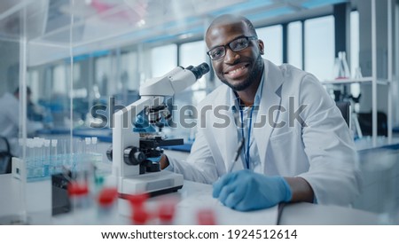 Modern Medical Research Laboratory: Portrait of Male Scientist Using Microscope, Charmingly Smiling on Camera. Advanced Scientific Lab for Medicine, Biotechnology, Microbiology Development Royalty-Free Stock Photo #1924512614