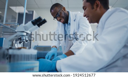 Modern Medical Research Laboratory: Two Smiling Male Scientists Working Together Using Microscope, Analysing Samples, Talking. Advanced Scientific Lab for Medicine, Biotechnology.