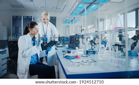 Modern Medical Research Laboratory: Two Female Scientists Working Using Digital Tablet, Analysing Biochemicals Samples. Scientific Lab for Medicine, Microbiology Development. Advanced Equipment Royalty-Free Stock Photo #1924512710