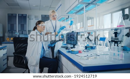 Modern Medical Research Laboratory: Two Female Scientists Working Using Digital Tablet, Analysing Biochemicals Samples. Scientific Lab for Medicine, Microbiology Development. Advanced Equipment Royalty-Free Stock Photo #1924512719