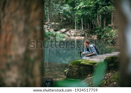 Woman photographer at a small mountain tropical lake. Young handsome caucasian girl tourist with a camera on a hike in a tropical forest