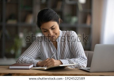 Happy millennial Indian woman sit at desk at home write study online on computer. Smiling young mixed race female work distant on laptop, take course on web. Education, learning concept. Royalty-Free Stock Photo #1924522589