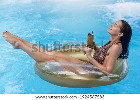 Happy relaxing woman swimming on float in pool, enjoying tropic cocktail, keeps eyes closed, sun tanning in swimming pool, wearing stylish swimsuit, summer vocation.