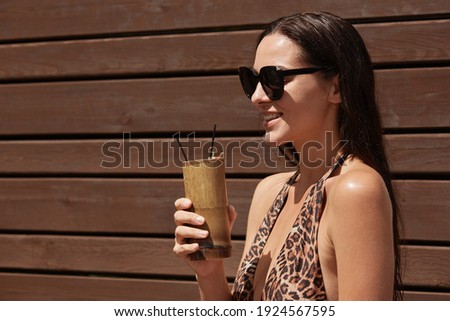 Woman with glass of cocktails posing profile against wooden wall, wearing stylish swimwear with leopard print and sunglasses, enjoying rest at sunny summer day.
