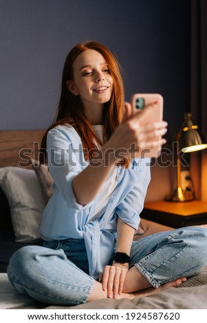 Charming redhead young woman taking mobile selfie photo on cellphone at cozy bedroom. Happy lady wearing home clothes making selfie on smartphone. Closeup portrait of cheerful female in bed.