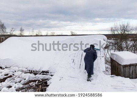 A man removes snow from the roof of a house and barn. A lot of snow on the roof of a rural house. A blizzard brought a lot of snow to the village. It is necessary to clean the snow on the roofs.