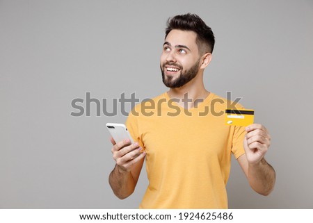 Young caucasian smiling bearded dreamful pensive student man 20s wear casual yellow basic t-shirt hold mobile cell phone credit bank card shopping online isolated on grey background studio portrait