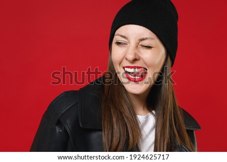 Close up of blinking young brunette woman 20s in basic casual black leather jacket white t-shirt hat showing making stick tongue out sign looking aside isolated on red background, studio portrait