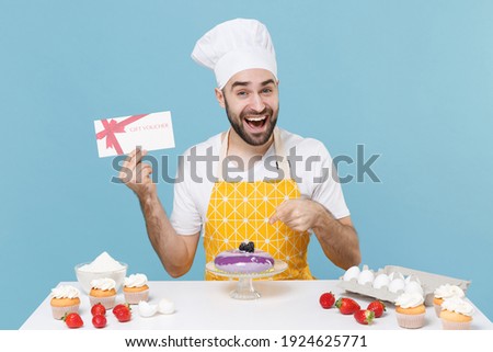 Funny young male chef or cook baker man in apron white t-shirt toque chefs hat cooking at table isolated on blue background. Cooking food concept. Hold gift certificate, pointing index finger on cake