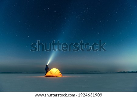 Tourist with flashlight near yellow tent lighted from the inside against the backdrop of incredible starry sky. Amazing night landscape. Tourists camp in snowy field. Travel concept