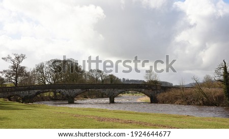 Stone Bridge over the river Ribble at Clitheroe Lancashire UK view across meadow to bridge with river in flood sweeping under historic bridge with rapids and bend in distance. Trees, winter sky.  