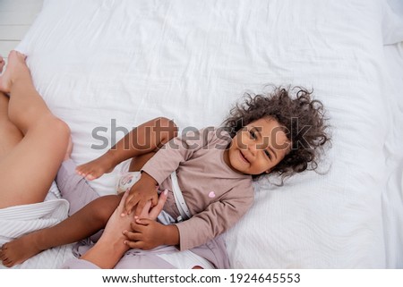 Young Caucasian blond mother tickles her little African American daughter. Lie on white bed, having fun, top view. The girls laugh, hug, the woman kisses the baby on the cheek. Home games, life style