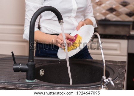 blonde in a white shirt washes dishes with a yellow washcloth in the kitchen.
