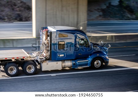 Big rig shiny blue semi truck tractor with long cab and auxiliary equipment on the back wall transporting flat bed semi trailer driving on the highway road under the concrete bridge Royalty-Free Stock Photo #1924650755