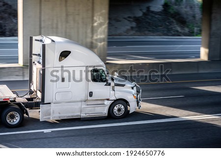 Big rig shiny white semi truck tractor with long cab and auxiliary equipment on the back wall transporting flat bed semi trailer driving on the highway road under the concrete bridge Royalty-Free Stock Photo #1924650776